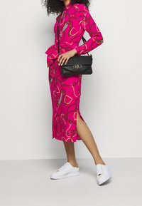 Kvinna iklädd en klänning i klar rosa med kedjemönster och midjeband, vita sneakers och håller en svart läderaxelväska.