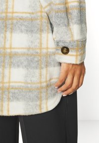 Camisa casaco em fleece com um padrão de xadrez em bege e cinza, apresentando uma bainha arredondada e um botão escuro no punho. Textura suave.