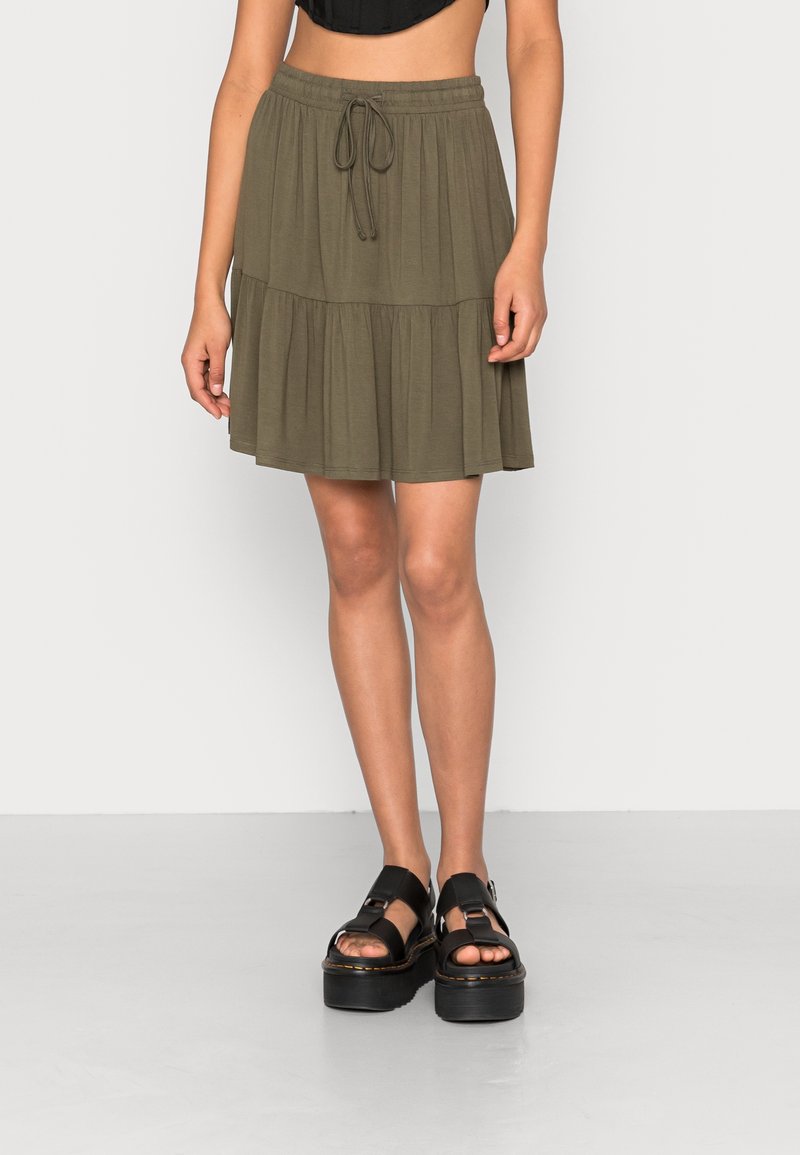 Mannequin féminin portant une mini-jupe à étages vert olive avec taille à cordon et sandales plateformes noires, debout devant un fond uni.