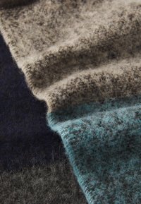 Wollen textiel met een zachte, fuzzy textuur. Kenmerkt zich door een kleurenverloop: beige, grijs en teal met duidelijke strengen die door elkaar zijn geweven.