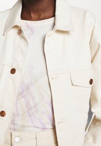 Kermel denimhansker med kobberknap-dekorationer, båret over en lys tie-dye skjorte med pastelfarvede lilla mønstre, parret med matchende kermel bukser.