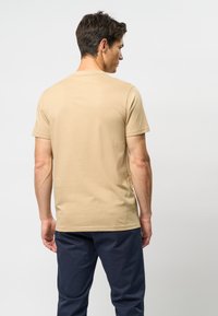 Beige bomulds t-shirt med korte ærmer, rund hals og en glat tekstur, båret af en person, der vender væk, kombineret med mørke bukser.