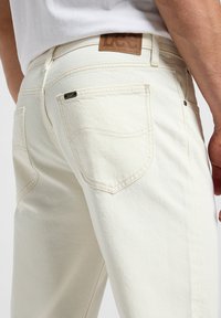 Jeans in denim bianco con una texture sottile, dotati di tasche posteriori e una patch in pelle marrone. Le cuciture e le suturazioni sono visibili.