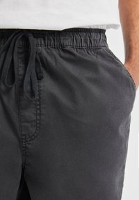 Pantalon de jogging noir avec une ceinture élastique, doté d'une fermeture à cordon de serrage et de poches latérales. Le tissu semble lisse et léger.