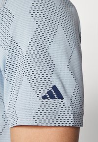 Chemise de sport bleu clair avec un motif texturé et un design géométrique bleu marine, avec un accent de logo bleu marine sur l'ourlet de la manche.