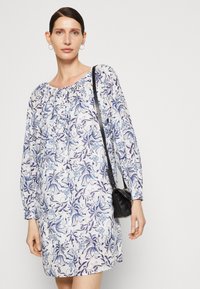 Trykt kjole med lange ermer, hvitt stoff med blå blomster mønster, og innsnøring ved halsen. En liten svart veske bæres på tvers av kroppen.