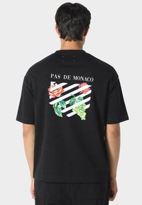 Czarna koszulka z nadrukiem z tyłu przedstawiającym róże i winogrona na białych ukośnych paskach, z napisem "PAS DE MONACO" nad wzorem.