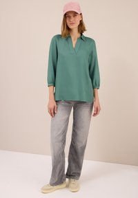 Frau mit einer rosa Mütze, einem grünen langärmeligen Blusenshirt, verwaschenen grauen Jeans und beigen Turnschuhen, die vor einem schlichten beigen Hintergrund steht.