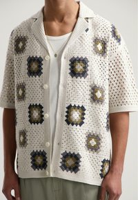 Open-knit haakshirt met korte mouwen, met een crèmekleurige basis en geruite vierkanten in donkerblauw, grijs en olijfgroen.