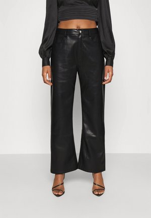 Femme portant un pantalon large taille haute en similicuir noir, un haut court noir et des sandales à talons noires à brides.