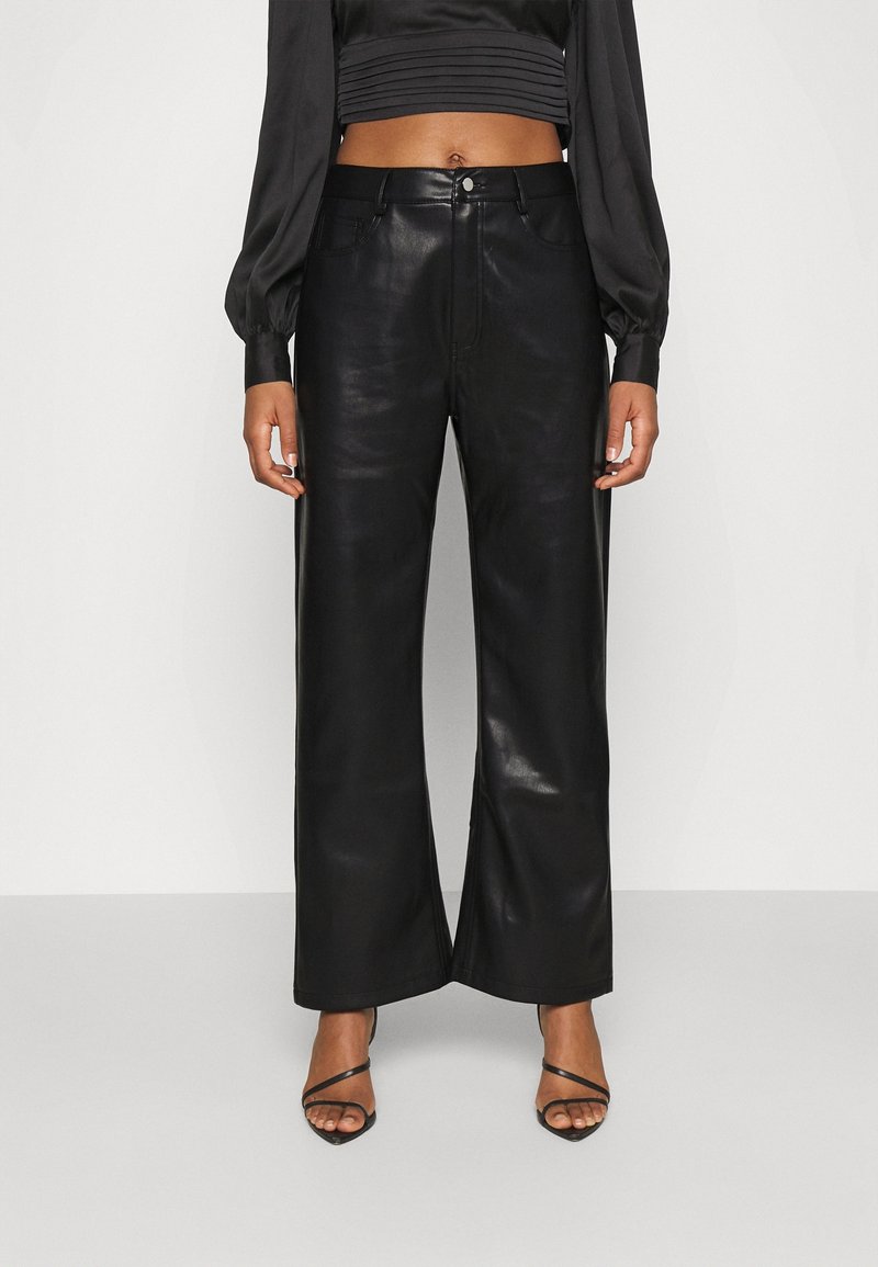 Femme portant un pantalon large taille haute en similicuir noir, un haut court noir et des sandales à talons noires à brides.