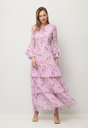 Kobieta ubrana w długą, różową sukienkę w kwiaty z warstwowymi falbanami i dzwonkowatymi rękawami, stojąca na tle jednolitego, jasnego tła.