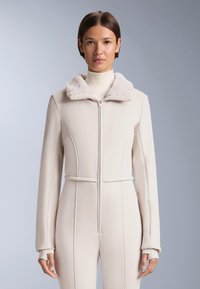 Kobieta ubrana w beżowy kombinezon na zamek z miękkim futrzanym kołnierzem i rękawami z otworem na kciuk, stojąca na jednolitym szarym tle.