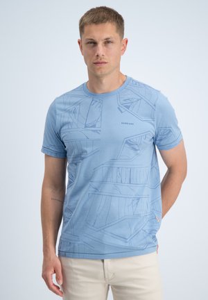 Man draagt lichtblauw grafisch T-shirt en beige broek, staande met één hand achter de rug tegen een effen achtergrond.