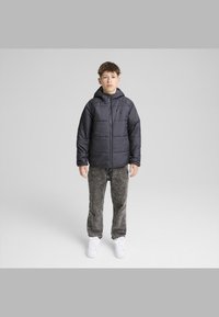 Svart puffjacka med huva, framdragkedja och en diskret logotyp. I kombination med grå strukturerade byxor och vita sneakers.