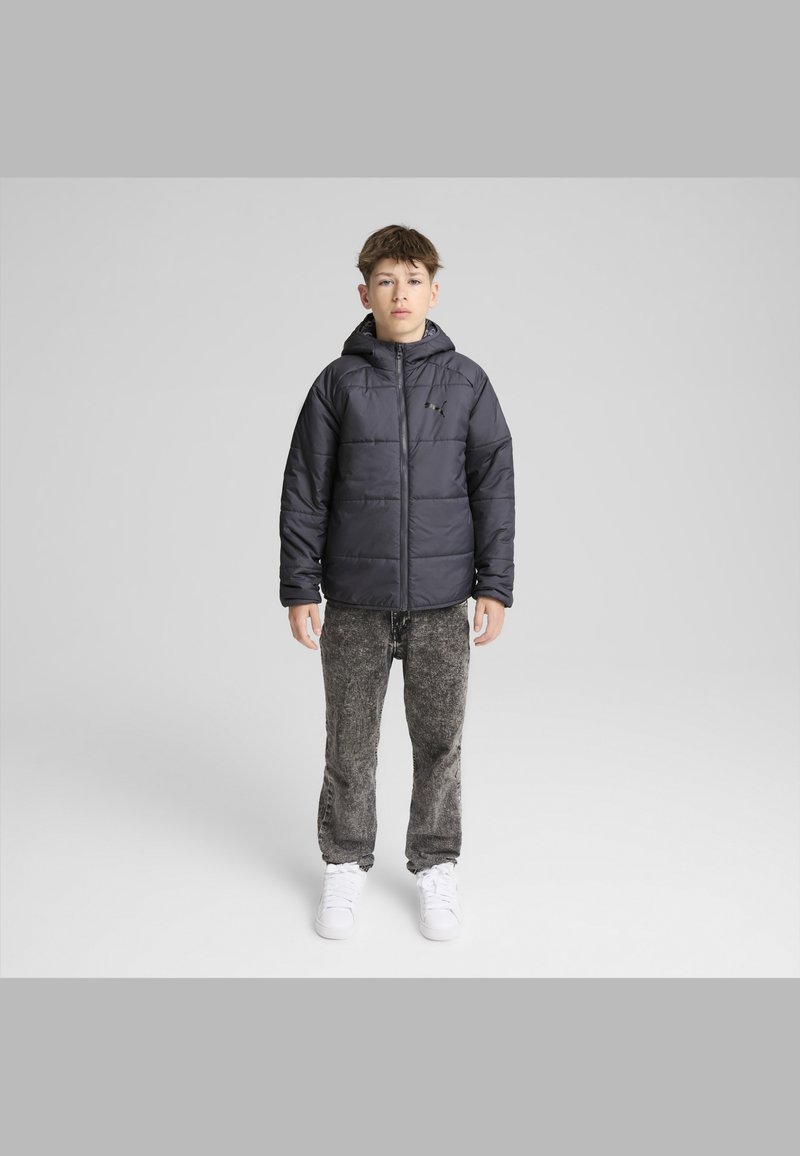 Svart puffjacka med huva, framdragkedja och en diskret logotyp. I kombination med grå strukturerade byxor och vita sneakers.