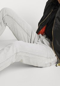 Persona vistiendo jeans gris claro y una chaqueta negra con cremallera dorada sentada en el suelo contra un fondo liso.
