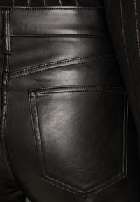 Pantaloni negri din piele cu o textură fină, cusături înclinate la yoke și detalii de cusătură subtile. Buzunarul din spate are o formă rectangulară.