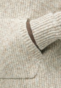 Μαλλινό πουλόβερ σε ανοιχτό μπεζ και κρέμα χρώμα, με ραβδωτή υφή και καφέ λεπτομέρεια στη διακόσμηση της τσέπης.