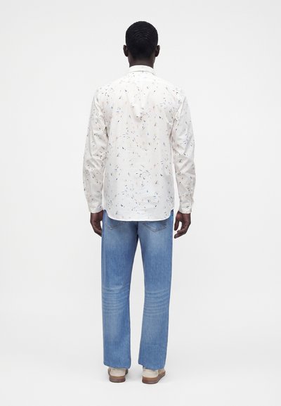Homme portant une chemise blanche à manches longues avec de petites taches colorées et un jean bleu, debout de dos devant un fond blanc uni.