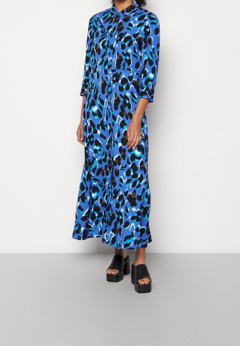 Femme portant une robe bleue à manches longues et longueur cheville avec un imprimé animal abstrait noir et turquoise, accompagnée de chaussures noires à plateforme et bout ouvert.