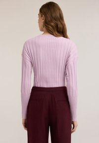 Maglione rosa chiaro a costine con maniche lunghe, indossato con pantaloni bordeaux scuro a vita alta. Texture morbida evidenziata da righe verticali.