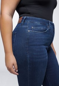 Middenhoge donkerblauwe denim jeans met een gladde structuur, voorzien van een bruine leren patch op de tailleband en twee kleine knoopjes op de voering van de voorkant.