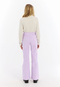 Lila skibroek met een flared ontwerp, featuring versterkte stiksels en een hoge tailleband. Gedragen met een ribbelige beige trui en zwarte laarzen.
