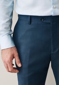 Man wearing light blue dress shirt tucked into dark blue tailored trousers with buttoned waistband and visible hand at side.