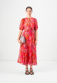 Woman wearing a red floral midi dress with puff sleeves, holding a light purple purse, and wearing black strappy sandals on a white background.