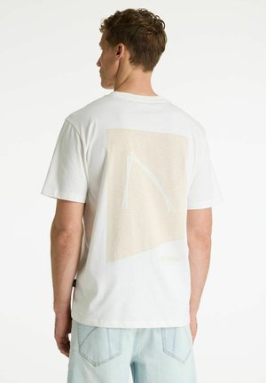 Jauns vīrietis ar īsiem matiem, ģērbies baltā T-kreklā ar gaiši bēšas abstraktas ģeometriskas apdrukas muguru un gaiši zilās džinsa biksēs.