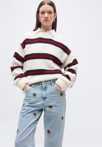 Γυναίκα που φοράει λευκό πουλόβερ με κόκκινες και μαύρες ρίγες πάνω από πουκάμισο με γιακά και ανοιχτόχρωμα τζιν με πολύχρωμα κεντημένα patches, με τα χέρια στις τσέπες.