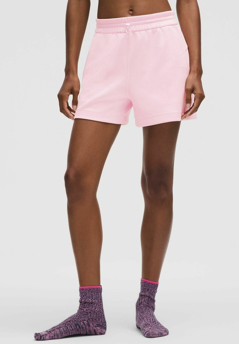 Rozā kokvilnas šortus ar elastīgu jostu un sānu kabatām apvieno ar teksturētām violētām zeķēm, kurām ir rozā aproce.