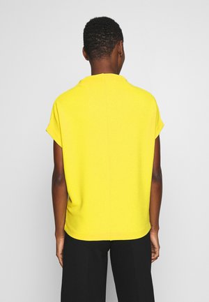 Osoba o krótkich kręconych włosach, ubrana w jasny żółty top z krótkim rękawem i czarne spodnie, stojąca tyłem na jednolitym tle.