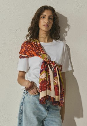 Meerjarige zijden sjaal gedrapeerd over een wit T-shirt, met bloemmotieven in rood, oranje en groen, en een gladde textuur.