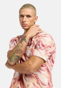 Kurze Hemdsärmel mit einem rosa, strukturierten Muster. Hat einen runden Ausschnitt und Bestickung auf der Brust. Tattoo am Arm sichtbar.