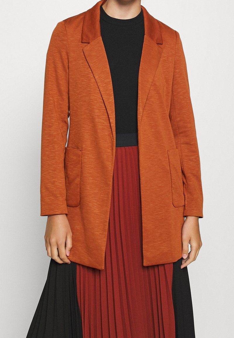 Rust-colored knit blazer with a smooth texture, notched lapels, and two front pockets, worn over a black top and red pleated skirt.
