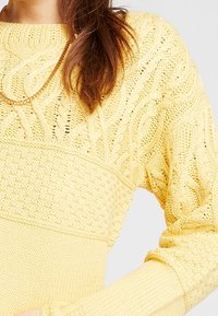 Pletený svetr v jemně žluté barvě s propracovanými copánkovými vzory, texturovanými panely a žebrovanými manžetami. Střední délka, lehký design.