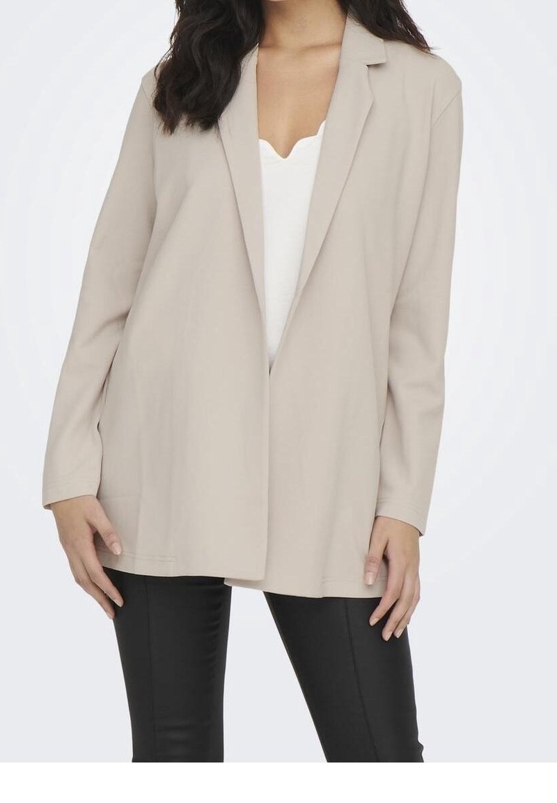 Femme portant un blazer beige ouvert sur un haut blanc et un pantalon noir slim, debout avec les mains détendues le long du corps.