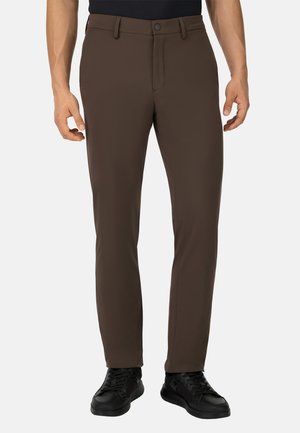 Hombre con pantalones rectos de color marrón oscuro y zapatillas negras, de pie con los brazos relajados a los lados, contra un fondo blanco.