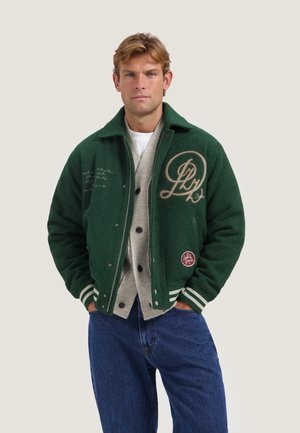 Jonge man met kort blond haar, gekleed in een groene varsityjas over een beige cardigan en blauwe spijkerbroek, handen in de zakken, neutrale achtergrond.