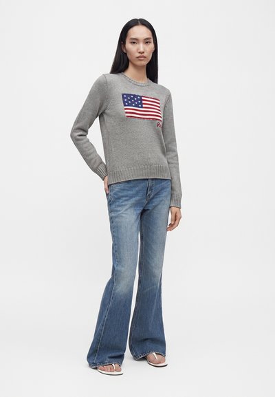 Femme debout portant un pull gris avec un motif du drapeau américain, un jean évasé bleu et des sandales blanches à bout ouvert sur fond blanc.