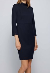 Námořnická modrá, sukňová šaty po kolena s vysokým límcem, dlouhými rukávy a bočními štěrbina. Vyrobeno z hladké, texturované látky s přiléhavým střihem.