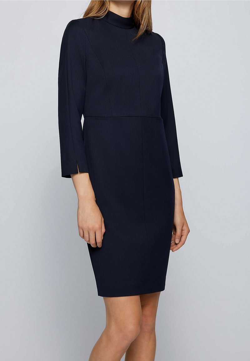 Námořnická modrá, sukňová šaty po kolena s vysokým límcem, dlouhými rukávy a bočními štěrbina. Vyrobeno z hladké, texturované látky s přiléhavým střihem.