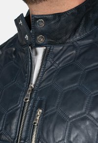 Navy leren jack met hexagonale stiksel, een hoge kraag met drukknoopsluitingen en zilveren ritsen aan de voorkant. Zachte textuur.