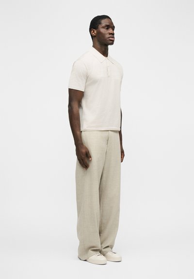 Άνδρας όρθιος, φοράει ανοιχτό μπεζ κοντομάνικο πόλο πουκάμισο, φαρδιά μπεζ παντελόνια και λευκά αθλητικά παπούτσια, σε απλό λευκό φόντο.