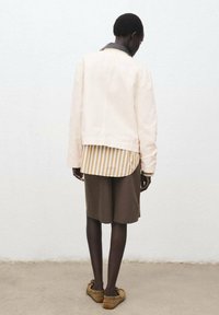 Light beige jacket with a black collar, layered over a striped shirt in beige and white, paired with knee-length brown shorts and brown shoes.
