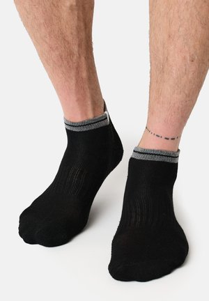 Ben iført sorte ankelsokker med grå kant, der viser en prikket linje-tatovering omkring højre ankel mod en ensfarvet baggrund.