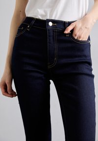 Jean denim bleu foncé à taille haute, avec surpiqûres jaunes contrastées, un bouton argenté, et des poches latérales. Texture lisse visible.