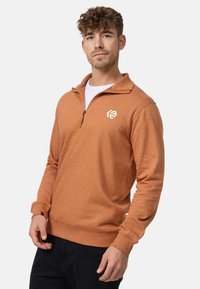 Orange zip-up sweatshirt med tekstureret finish, der har en ståkrave og et broderet logo på venstre bryst. Lange ærmer.